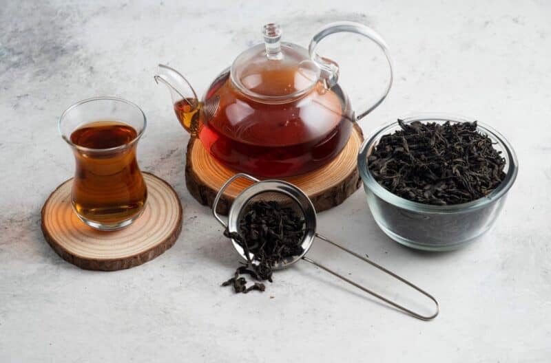 Black tea in a glass tea cup and glass tea pot with a bowl of black tea leaves